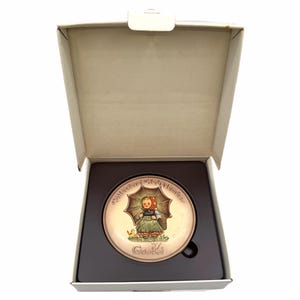 May include: A round Goebel Collector's Club Member plate featuring a colorful illustration of a child holding an umbrella and a basket, presented in a square cardboard box with a dark brown molded insert to hold the plate securely in place.