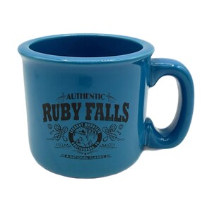 Könnte beinhalten: Eine blaue Keramik-Tasse mit gebogenem Henkel. Die Tasse trägt den schwarzen Schriftzug "AUTHENTIC RUBY FALLS", ein rundes Grafikdesign und die Worte "A NATIONAL CLASSIC". Die Tasse ist für Getränke konzipiert.