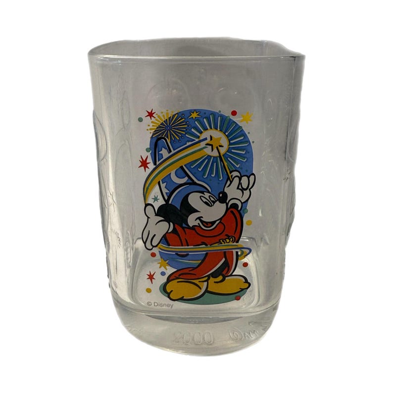 May include: Clear glass tumbler featuring a colorful graphic of Mickey Mouse in a sorcerer's outfit, with a blue and yellow background. The tumbler has the year 2000 printed on the bottom. The Disney logo is also visible.