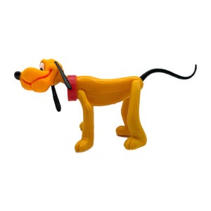 Könnte beinhalten: Gelbe Pluto-Figur mit schwarzem Schwanz und Ohren, rotem Halsband und schwarzer Nase. Der Hund steht, blickt nach links und lächelt leicht.