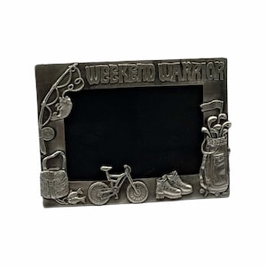 May include: A rectangular silver-toned metal picture frame featuring the embossed text WEEKEND WARRIOR at the top. The frame is decorated with raised motifs including a fishing rod, a bicycle, hiking boots, and a golf bag with clubs.