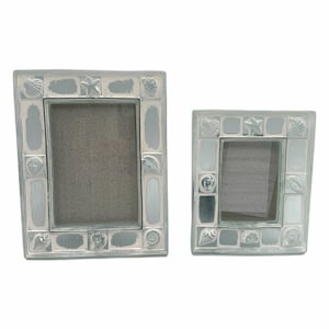 May include: Two silver-colored picture frames with a coastal theme. The frames are decorated with seashells and starfish. The frames have a rectangular shape with a light blue and white painted border.