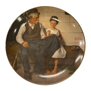 Op de afbeelding: Decoratieve plaat met een schilderij van Norman Rockwell. Het kunstwerk toont een oudere man en een jongere die op een bank zitten te naaien. De plaat heeft een gouden rand en een warm kleurenpalet.