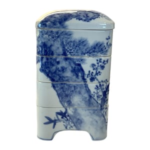 Könnte beinhalten: Eine blau-weiße Bento-Box aus Keramik mit Deckel. Die Box ist rechteckig mit abgerundeten Ecken und zeigt ein gemaltes Baum- und Blumendesign. Die Box ist in vier Abschnitten gestapelt und hat einen kleinen Fuß.