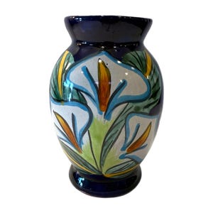 Puede incluir: Un jarrón de cerámica con borde y base azul oscuro, con un diseño floral pintado a mano. Las flores son blancas con detalles azules, amarillos y naranjas, y hojas verdes. El jarrón mide aproximadamente 20 cm de altura.