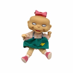 Puede incluir: Muñeca de un personaje de bebé con un lazo rosa, que lleva una camisa rosa a rayas, una falda verde con un pato amarillo y zapatos rosas. La muñeca tiene una expresión sonriente.