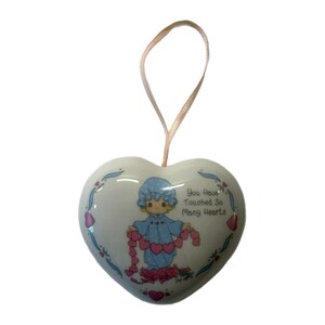 Könnte beinhalten: Herzförmiger Keramikschmuck mit hellblauem Band. Der Schmuck zeigt eine Cartoonfigur in einem blauen Kleid, die eine Girlande aus rosa Herzen hält. Der Text lautet "You Have Touched So Many Hearts."