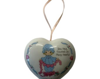 Puede incluir: Adorno de cerámica en forma de corazón con una cinta azul claro. El adorno presenta una figura de dibujos animados con un vestido azul que sostiene una guirnalda de corazones rosas. El texto dice "You Have Touched So Many Hearts."