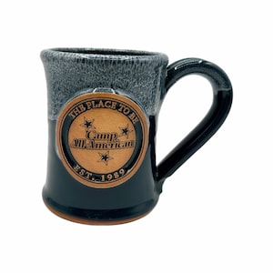 Könnte beinhalten: Eine Keramik-Tasse mit dunkelblauem Boden und gesprenkeltem grauem Oberteil. Die Tasse hat ein bronzefarbenes Emblem mit dem Text "THE PLACE TO BE Camp All-American EST. 1989" und Stern-Grafiken. Der Griff ist schwarz.