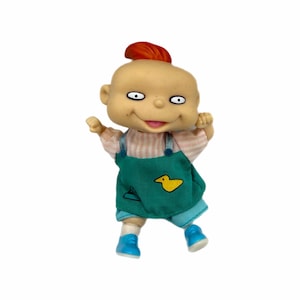 Puede incluir: Muñeca de plástico de un personaje de dibujos animados con un mechón de pelo rojo. La muñeca tiene una cara bronceada con ojos grandes y una expresión sonriente. Viste una camisa a rayas rosas y blancas, un peto verde y zapatos azules. Un pato amarillo está en el peto.