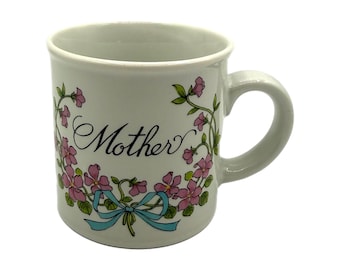 Könnte beinhalten: Weiße Keramiktasse mit dem Wort "Mother" in Schreibschrift, umgeben von rosa Blumen und einer hellblauen Schleife. Die Tasse hat einen gebogenen Henkel und ein klassisches Design.