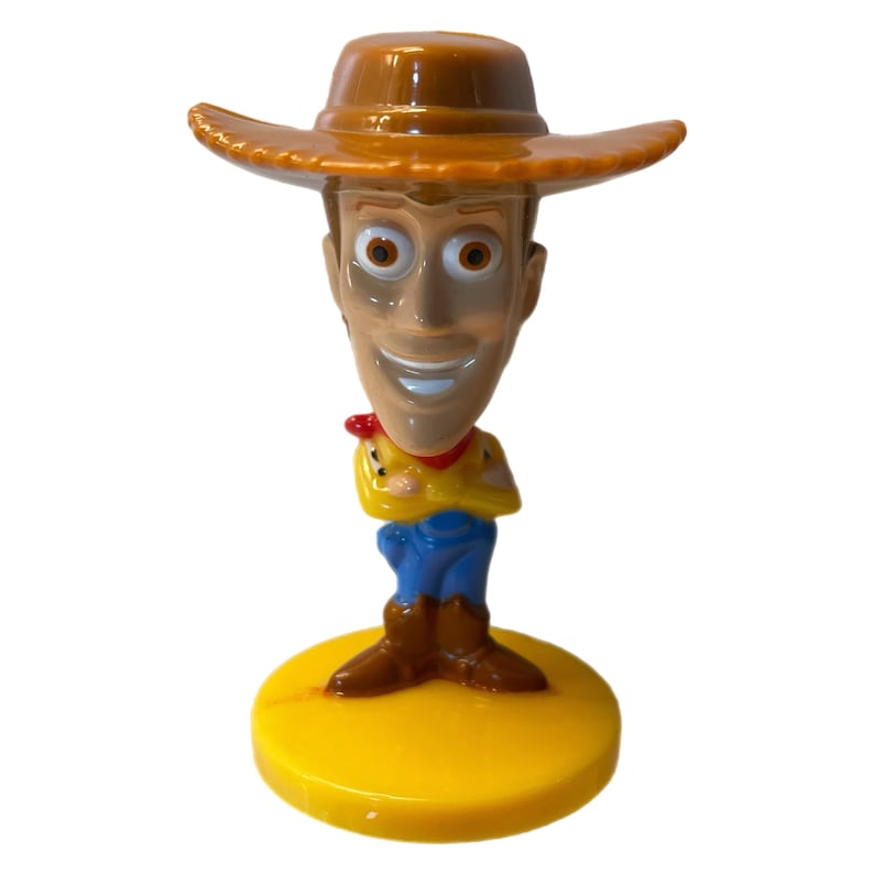 Może przedstawiać: Plastikowa figurka Woody'ego z Toy Story. Woody ma na sobie sw&oacute;j charakterystyczny brązowy kowbojski kapelusz, niebieskie dżinsy i ż&oacute;łtą kamizelkę. Uśmiecha się i ma brązowe oczy. Figurka stoi na ż&oacute;łtej podstawie.
