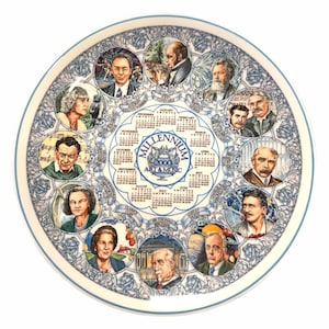 May include: A decorative plate featuring portraits of historical figures and a calendar for the year 2000. The plate has a white background with a blue floral border and a central design that reads "Millennium 2000 Art & Music."