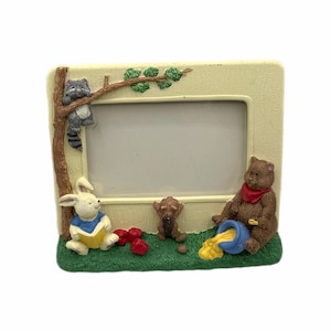 May include: A rectangular picture frame with a light yellow border and a green base. The frame is decorated with cartoon animals, including a raccoon, a rabbit, a monkey, and a bear. The bear is pouring honey from a blue pot.
