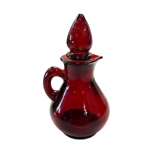 May include: A ruby red glass cruet with a handle and a spout. The cruet has a bulbous body and a tall, pointed stopper. The glass is a deep, transparent red, and the cruet is likely vintage or antique.