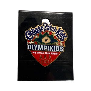 May include: A Cabbage Patch Kids OlympiKids 1996 USA pin. The pin is a shield shape with a red background and gold trim. The pin features the Cabbage Patch Kids logo, the text "OLYMPIKIDS", and the Olympic rings.