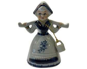 May include: A ceramic figurine of a woman in traditional Dutch attire. The figurine is white with blue accents and gold trim. It features a bonnet, dress, and a small bucket. The dress has a floral design.