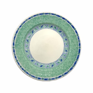 May include: A round ceramic plate with a white center and a green and blue patterned border. The border features a speckled green background with blue and green leaf and floral designs. The outer rim is a solid blue with small floral accents.