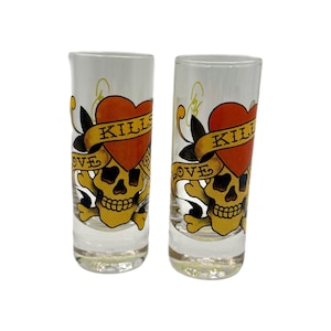 Puede incluir: Dos vasos de chupito transparentes con un diseño de calavera y huesos cruzados. El diseño presenta una calavera amarilla con un corazón rojo, una pancarta que dice "Kills Love" y detalles en negro. Los vasos son cilíndricos.