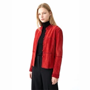May include: A red suede jacket with a round collar and button fastening. The jacket has a patchwork design with visible stitching and a knitted trim on the cuffs. The model is wearing a black turtleneck and black trousers.