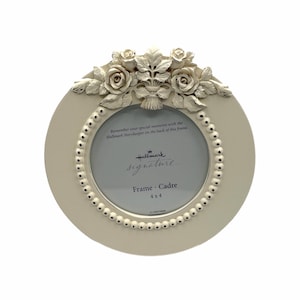 May include: A cream-coloured round picture frame with a floral rose design at the top. The frame has a beaded border and holds a 10x10 cm photo. Text on the frame reads "Hallmark signature" and "Frame Cadre".
