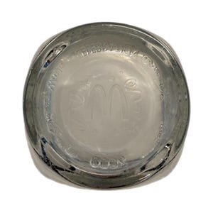 May include: Clear glass McDonald's commemorative drinking glass. The bottom of the glass features the McDonald's logo and text. The glass has a rounded square shape and is slightly tinted.
