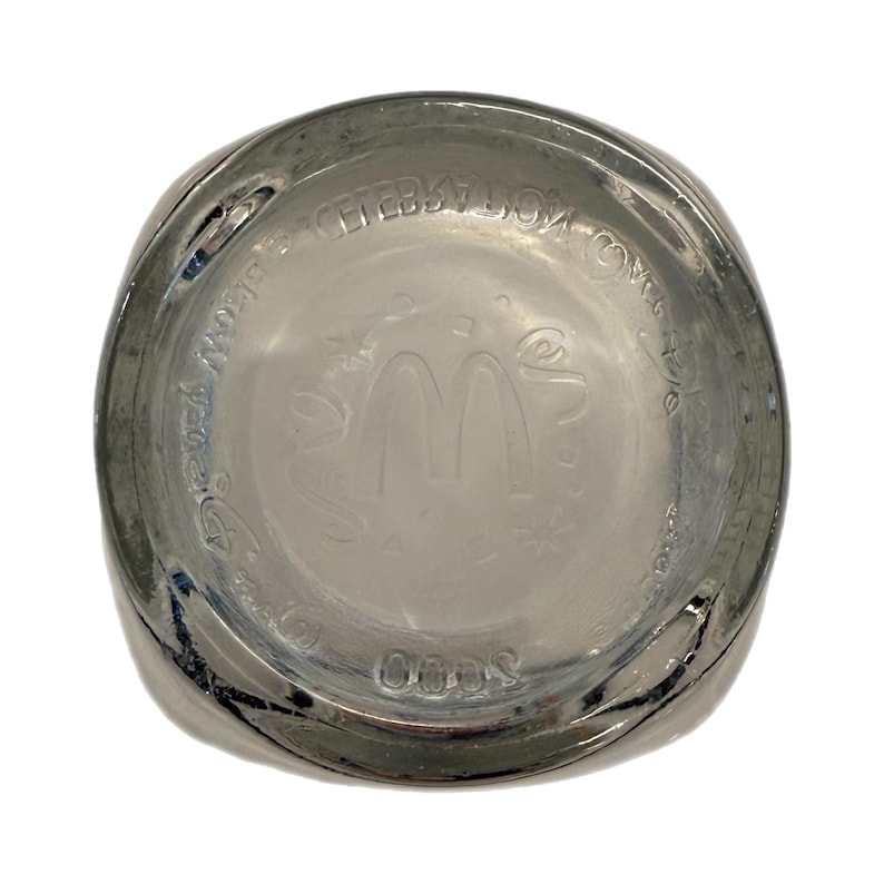 May include: Clear glass McDonald's commemorative drinking glass. The bottom of the glass features the McDonald's logo and text. The glass has a rounded square shape and is slightly tinted.