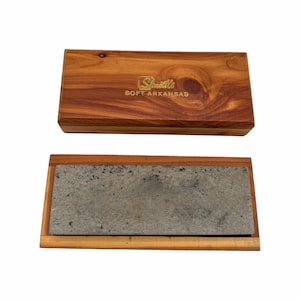 May include: A rectangular sharpening stone set in a wooden base with a matching wooden lid. The lid features the gold-colored text Smith's SOFT ARKANSAS. The stone is light gray with dark speckles, and the wooden box has a natural wood grain finish.