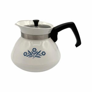 Puede incluir: Cafetera vintage blanca con asa y tapa negras. La cafetera presenta un diseño floral azul y una banda plateada en el centro. El pico es blanco y el asa es curva.