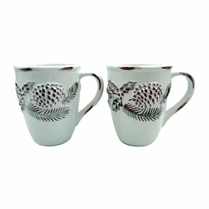 May include: Two white ceramic mugs with a rustic design. Each mug features a raised relief of a pinecone and foliage in shades of brown and gray. The rims and handles have a brown trim, adding to the vintage aesthetic. Perfect for serving hot beverages.