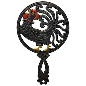 May include: Black cast iron trivet featuring a rooster design. The rooster is within a circular frame with a decorative handle. The rooster has red, yellow, and black accents.