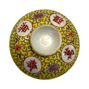 May include: A yellow ceramic lid with a central white cup. The lid features red Chinese characters in white circles, pink flowers, and a swirling pattern. The lid is likely for a tea cup or small bowl.