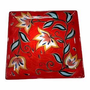 May include: A square ceramic plate with a vibrant red background featuring a hand-painted floral pattern. The design includes three large stylized flowers in shades of yellow, white, and orange, accented by dark blue and light blue leaves and vines.