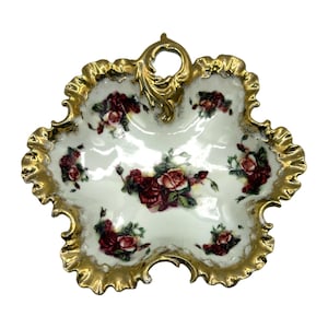 May include: A decorative, white porcelain dish with a scalloped, gold-colored rim. The dish features a floral pattern of red roses and green leaves. A gold handle is at the top. The dish is shaped like a six-petal flower.