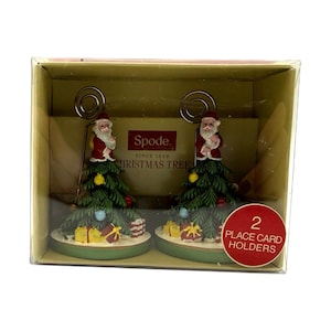Op de afbeelding: Twee kerstboom-naamkaarthouders in een doorzichtige doos. Elke houder heeft een groene boom met ornamenten en een kerstmanfiguur. De doos heeft een rode sticker met de tekst "2 Naamkaarthouders". De merknaam "Spode" is zichtbaar.