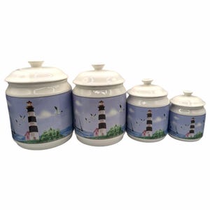 May include: A set of four white ceramic canisters with lids, arranged in descending order of size. Each canister features a wrap-around illustration of a black and white lighthouse, green coastal foliage, and seagulls against a blue sky.