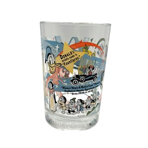 Puede incluir: Vaso transparente con personajes de Disney, incluyendo el Pato Donald, Blancanieves y los Siete Enanitos. El vaso tiene texto que dice "Donald's middle name is Fauntleroy" y "Disney Stars & Motor Cars Parade features classic American cars from 1929 to 1951."