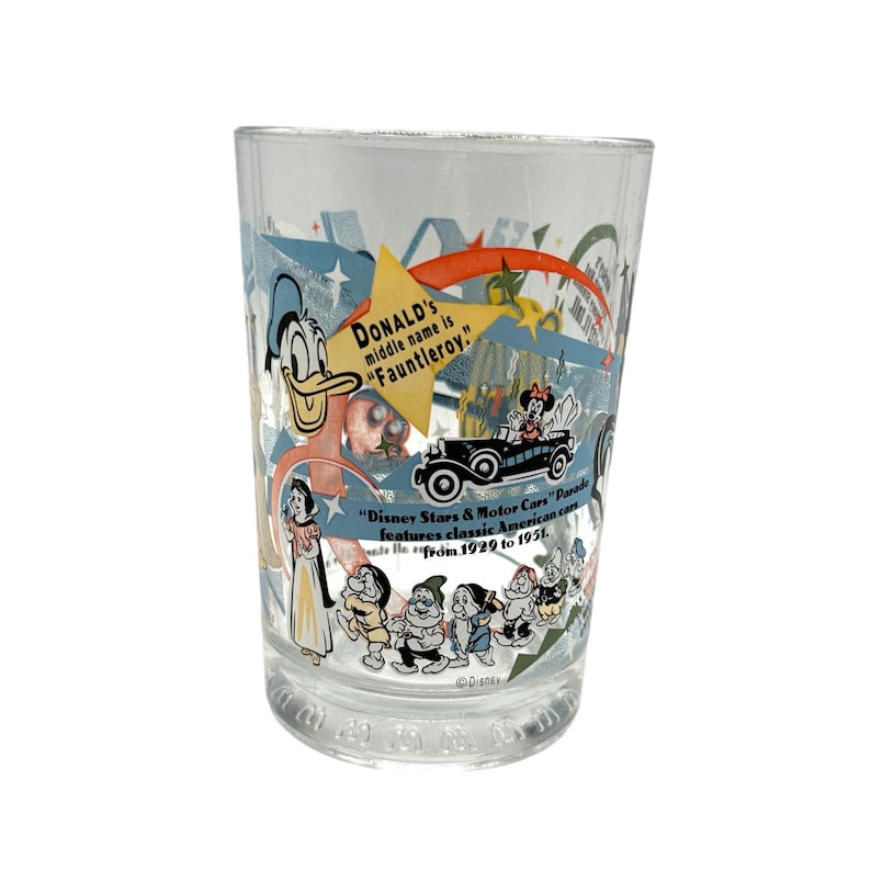 Puede incluir: Vaso transparente con personajes de Disney, incluyendo el Pato Donald, Blancanieves y los Siete Enanitos. El vaso tiene texto que dice "Donald's middle name is Fauntleroy" y "Disney Stars & Motor Cars Parade features classic American cars from 1929 to 1951."