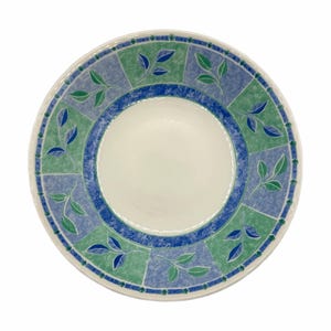 Op de afbeelding: Een ronde, witte keramische plaat met een decoratieve rand. De rand heeft een blauw en groen geblokt patroon met bladaccenten. Een blauwe ring omcirkelt het midden en de plaat heeft een wit interieur.