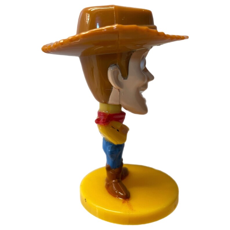 Może przedstawiać: Plastikowa figurka Woody'ego z Toy Story. Woody ma na sobie sw&oacute;j charakterystyczny brązowy kowbojski kapelusz, czerwoną chustkę i niebieskie dżinsy. Stoi na ż&oacute;łtej podstawie.