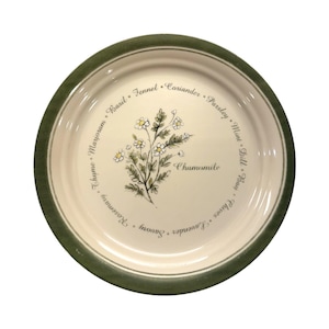 Peut inclure: Une assiette ronde en céramique blanc cassé avec un bord vert. L'assiette présente une illustration florale de camomille et les noms d'herbes et d'épices, dont le basilic, le fenouil et la coriandre, sur le bord.