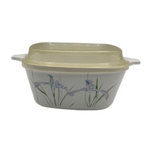 Pode incluir: Uma travessa retangular, branco-sujo, com tampa amarelo-pálido. A travessa apresenta um desenho floral de íris roxas e folhas verdes. A tampa tem uma forma ligeiramente curva e encaixa bem.