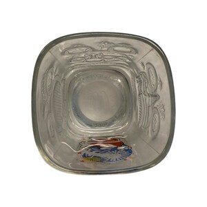 May include: Clear glass tumbler with a square base and rounded corners. The glass features embossed designs on the sides and a colorful emblem at the bottom. The interior has a circular design with the letter "M".