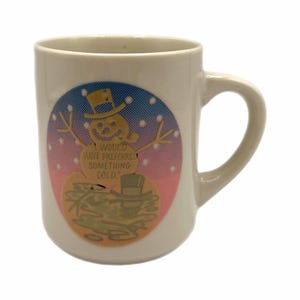 May include: Cream-colored ceramic mug with a whimsical snowman graphic. The snowman wears a top hat and has outstretched arms. The mug features the text: "I WOULD HAVE PREFERRED SOMETHING COLD." The background is a gradient of pink and blue.