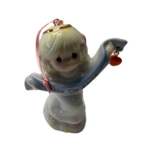 May include: A ceramic angel figurine with a light blue robe and blonde hair. The angel holds a banner that reads "Peace On Earth" and a small red heart charm. A pink ribbon is attached for hanging.