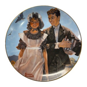 Op de afbeelding: Decoratieve bord met een geschilderde scène van een jong stel dat buiten loopt. Het bord heeft een gouden rand en een blauwe luchtachtergrond met vogels. Het kunstwerk is linksonder gesigneerd met "Norman Rockwell".