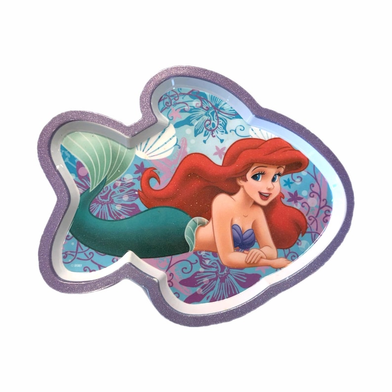 May include: A fish-shaped tray featuring Ariel from The Little Mermaid. The tray has a purple glittery border and a colourful interior with Ariel's image. Ariel has red hair, a blue top, and a green tail. The background has floral patterns.