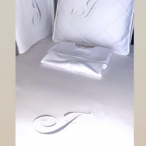 May include: White baby bedding set featuring a quilted pillow and a white onesie. The pillow and onesie are embroidered with a cursive letter "J".