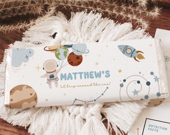 May include: A white chocolate bar wrapper with a space theme. It features an astronaut holding balloons with planets, a rocket ship, and stars. The text on the wrapper says "MATTHEW'S 1st trip around the sun!"
