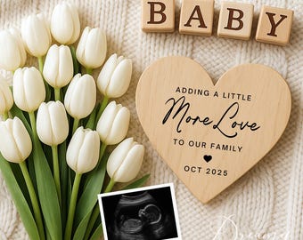 May include: Wooden heart-shaped sign with the text "Adding a little More Love to our Family" and the date "Oct 2025".  Four wooden blocks spelling "BABY" are on a white knitted surface with a bouquet of white tulips and a black and white ultrasound image.